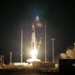 Ночной запуск ракеты в космос с освещенной площадки и дымом на заднем плане.