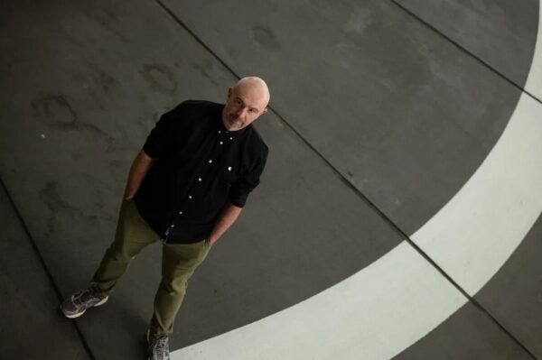 Человек в черной рубашке и зеленых брюках стоит на сером фоне с белой полосой.