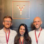 Три человека с бейджами на фоне логотипа Y на темной деревянной стене.