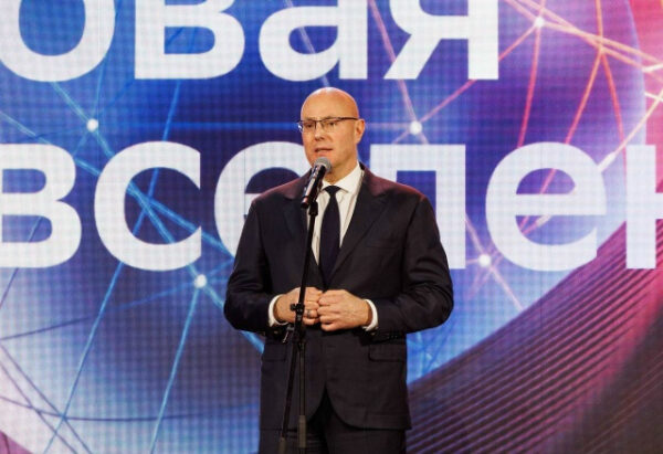 Мужчина в костюме с микрофоном на фоне экранной надписи на мероприятии.