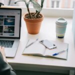 Рабочее место у окна: ноутбук, блокнот, смартфон, ручка и растение в горшке.