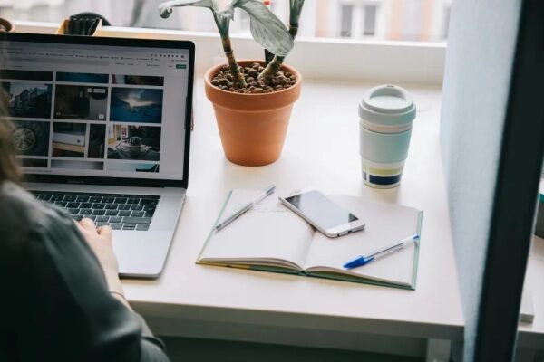 Рабочее место у окна: ноутбук, блокнот, смартфон, ручка и растение в горшке.