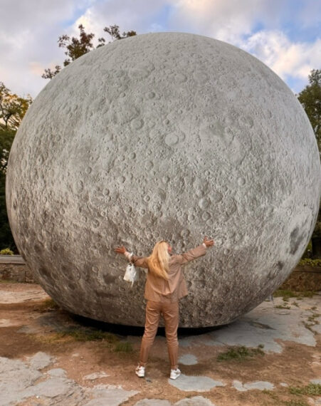 Человек обнимает модель луны на улице, ясное небо с облаками сверху.