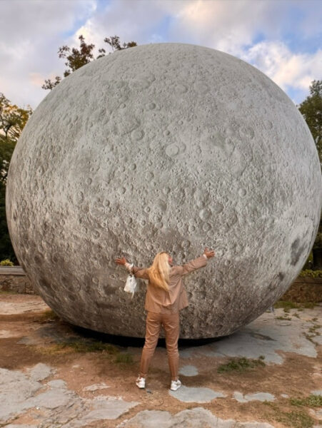 Человек обнимает модель луны на улице, ясное небо с облаками сверху.