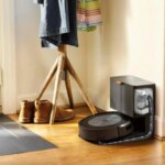 Робот-пылесос на базе, рядом обувь и вешалка на деревянном полу в светлой комнате.