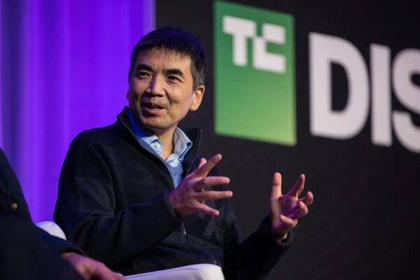 Мужчина на конференции TechCrunch Disrupt, жесты, темный фон.