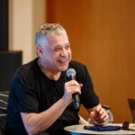 Мужчина в черной футболке выступает с микрофоном, сидя за круглым столом.