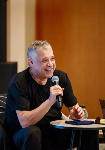 Мужчина в черной футболке выступает с микрофоном, сидя за круглым столом.