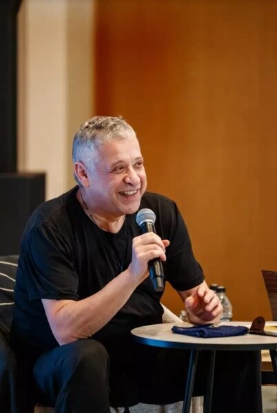 Мужчина в черной футболке выступает с микрофоном, сидя за круглым столом.