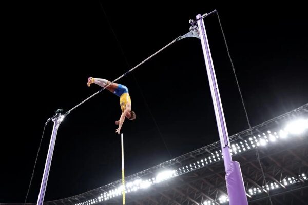 Спортсмен прыгает с шестом на соревнованиях ночью, стадион освещён прожекторами.