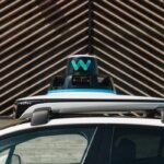 Автомобиль Waymo с лидаром на крыше у геометрической стены.