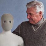 Старший мужчина обнимает робота с нейтральным лицом на сером фоне.