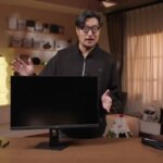 Человек демонстрирует монитор на столе в уютной комнате с мягким освещением.