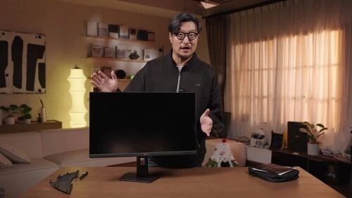 Человек демонстрирует монитор на столе в уютной комнате с мягким освещением.