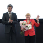 Чествование: женщина с букетом цветов и медалями принимает поздравления на сцене.