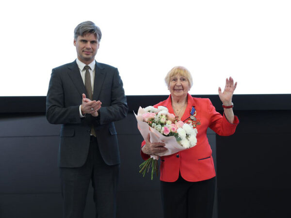 Чествование: женщина с букетом цветов и медалями принимает поздравления на сцене.