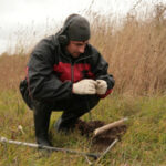 Человек с металлоискателем и лопатой исследует землю в поле осенью.