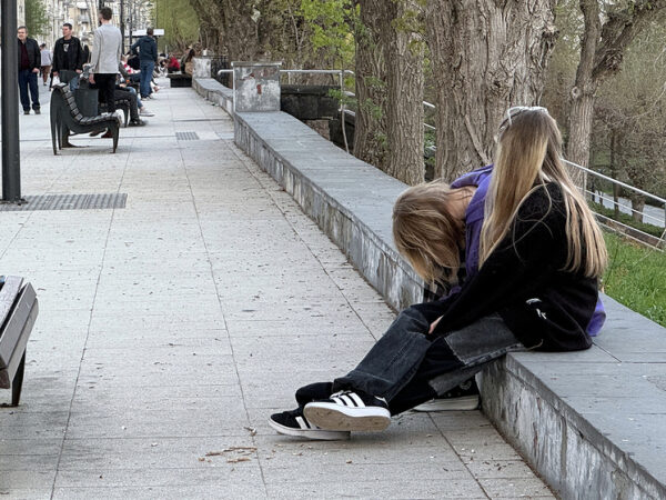 Две девушки сидят на набережной, согнувшись, на фоне деревьев и скамейки.