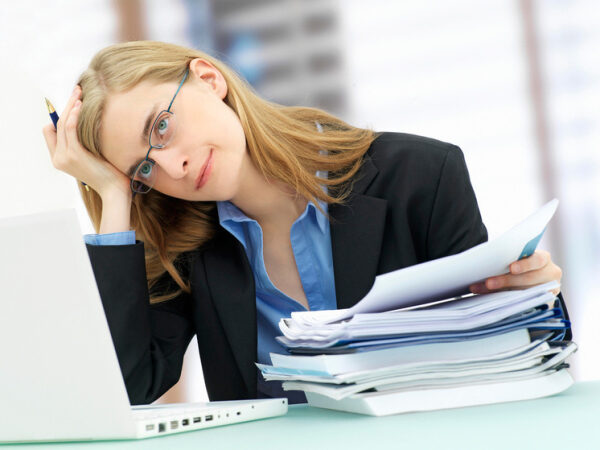 Женщина за ноутбуком с документами, выражение усталости; офисная работа.