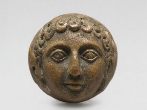 Древняя бронзовая скульптура лица с вьющимися волосами на светлом фоне.