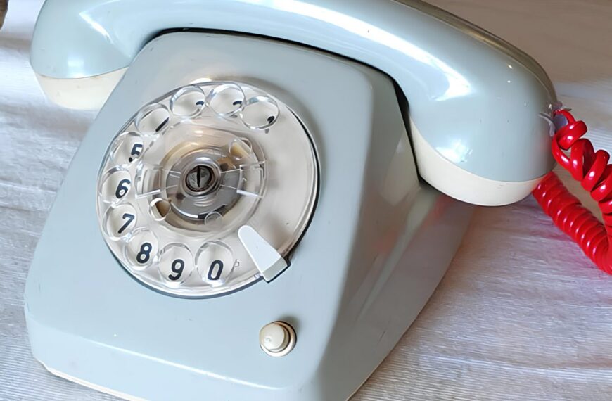 Старый дисковый телефон с красной трубкой на белой поверхности.