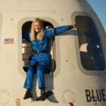 Человек в синем комбинезоне сидит в люке капсулы Blue Origin, руки разведены в стороны.