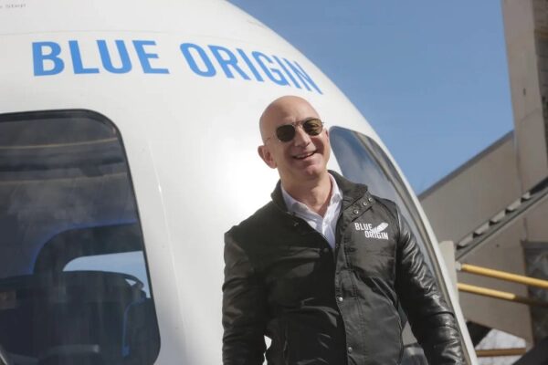 Человек в куртке Blue Origin у капсулы Blue Origin на фоне здания и голубого неба.