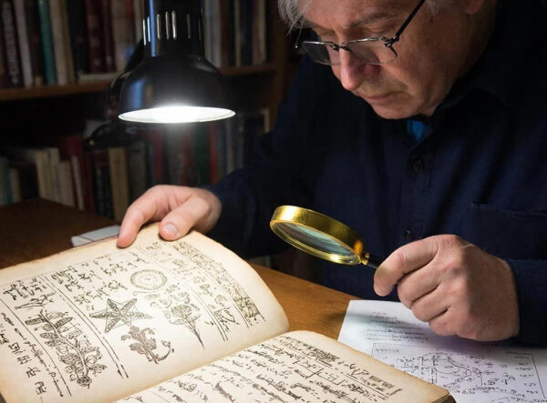 Человек изучает старинную книгу с лупой под лампой в библиотеке.