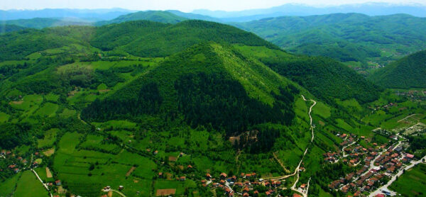 Зеленый холм пирамидальной формы рядом с деревней и горными пейзажами.