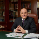 Человек в деловом костюме сидит за большим столом в кабинете с книгами.