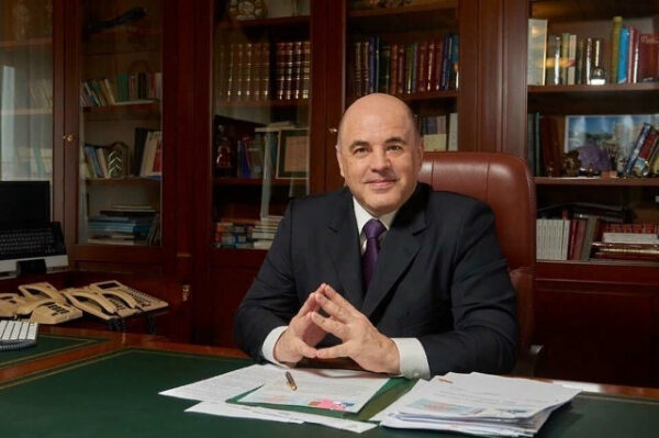 Человек в деловом костюме сидит за большим столом в кабинете с книгами.