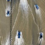 Автомобили едут по затопленной дороге после наводнения, создавая волны на воде.