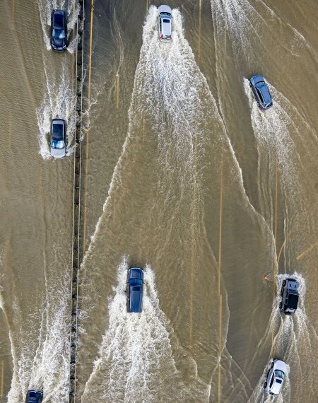 Автомобили едут по затопленной дороге после наводнения, создавая волны на воде.