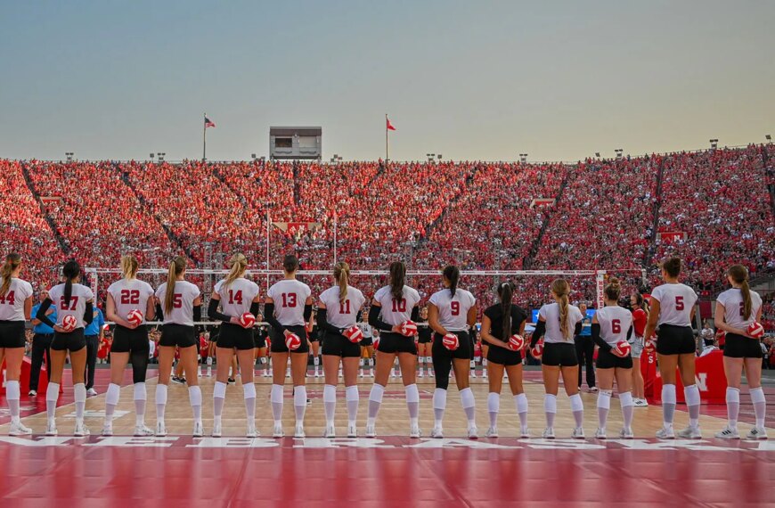 Волейбольная команда перед матчем на стадионе, заполненном зрителями в красной одежде.