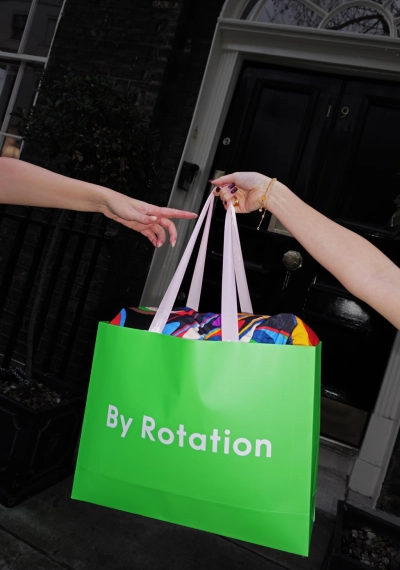 Две руки держат зеленый шопинг-пакет с одеждой перед черной дверью.