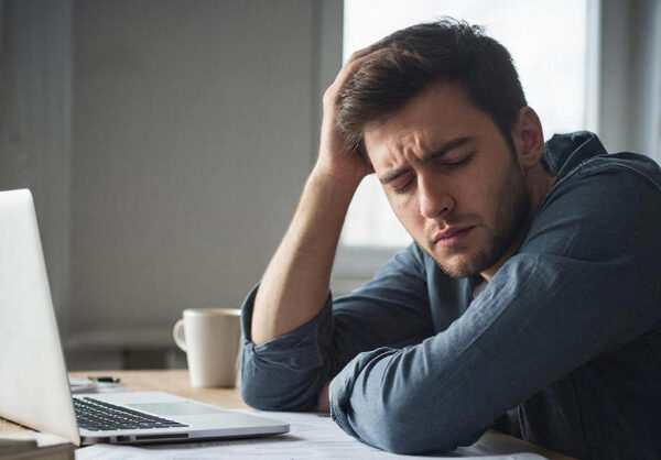 Мужчина за ноутбуком выглядит усталым и задумчивым, сидя за столом с бумагами и кружкой.