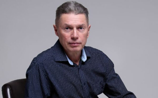 Мужчина в темной рубашке сидит на стуле, серый фон.