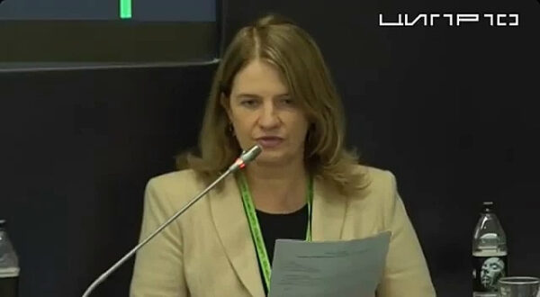 Женщина читает документ на конференции, рядом с ней микрофон и бутылка воды.