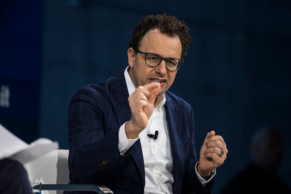 Мужчина в очках и костюме выступает на конференции, жестикулируя рукой.