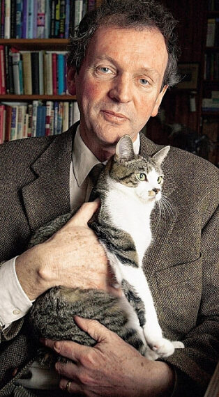 Мужчина в костюме держит полосатую кошку на фоне книжных полок.