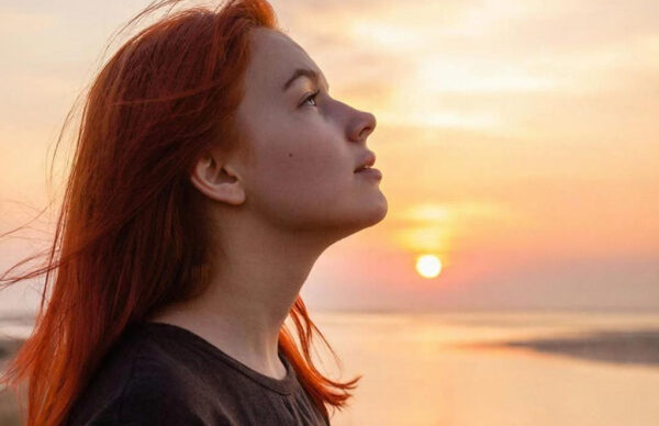 Женщина с рыжими волосами смотрит на закат у моря.