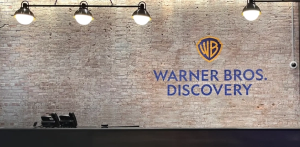 Стенд с логотипом Warner Bros. Discovery на кирпичной стене, светильники сверху.