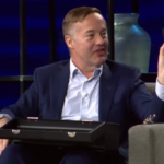 Мужчина в костюме держит черный ящик и небольшой пакет в студии, сидя в кресле.