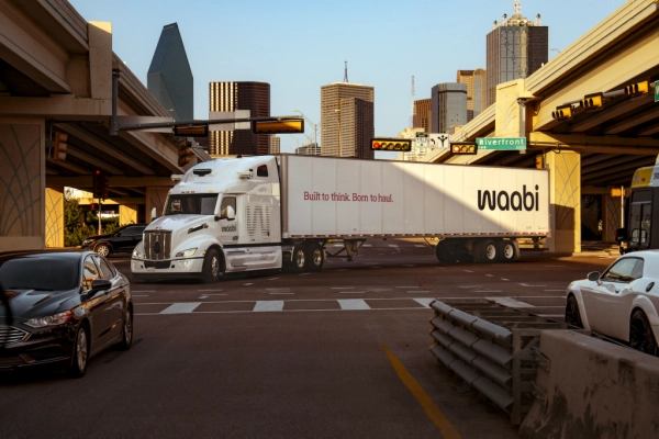 Белый грузовик Waabi едет по городскому перекрестку с множеством автомобилей.