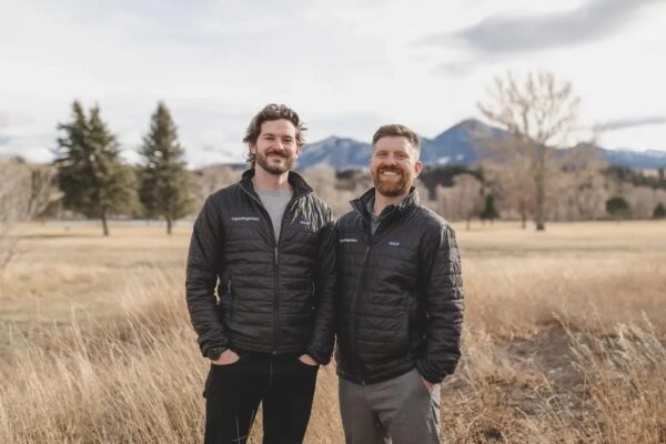 Два улыбающихся человека на природе в куртках, на фоне горы и деревья.