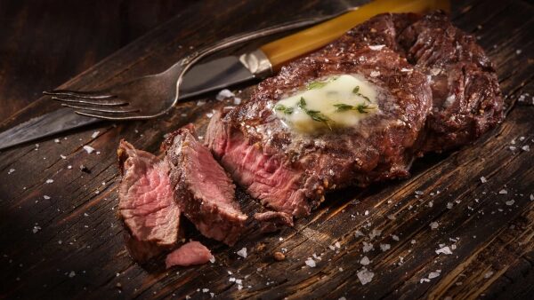 Сочный стейк с маслом и розмарином на деревянной доске, рядом вилка и нож.