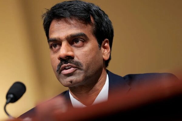 Мужчина с усами за микрофоном на фоне золотистого оттенка, говорящий серьезно.