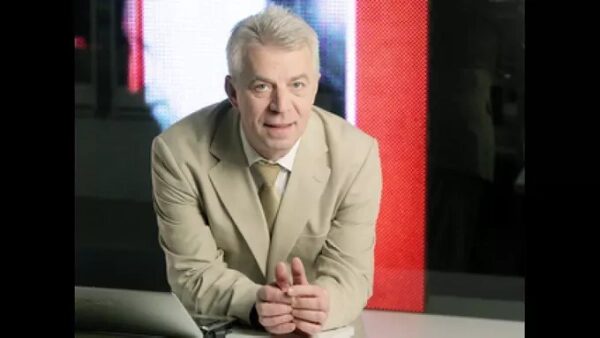 Мужчина в светлом костюме стоит, слегка опираясь на стол, фон с экраном.