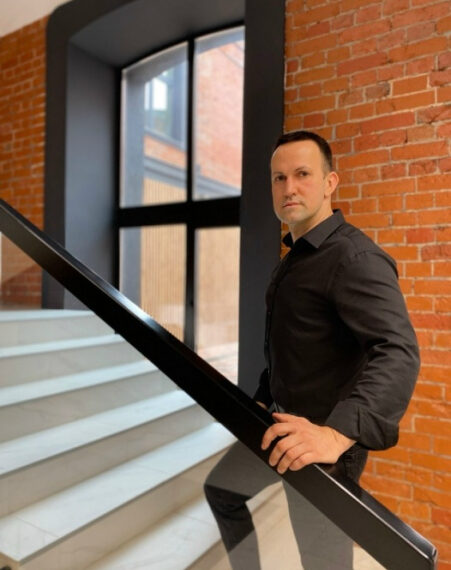 Мужчина в черной рубашке на лестнице с кирпичной стеной на фоне.