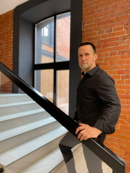 Мужчина в черной рубашке на лестнице с кирпичной стеной на фоне.